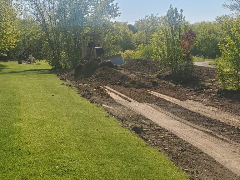 A construction vehicle clears dirt on a path through a grassy field bordered by trees on a sunny day.
