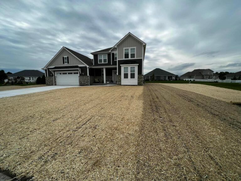 A two-story suburban house with a large, newly seeded front lawn covered in straw mulch under a cloudy sky.