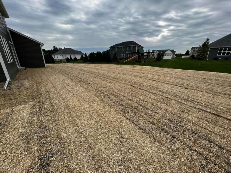 A freshly seeded backyard covered in straw mulch next to a modern dark-sided house under a cloudy sky.