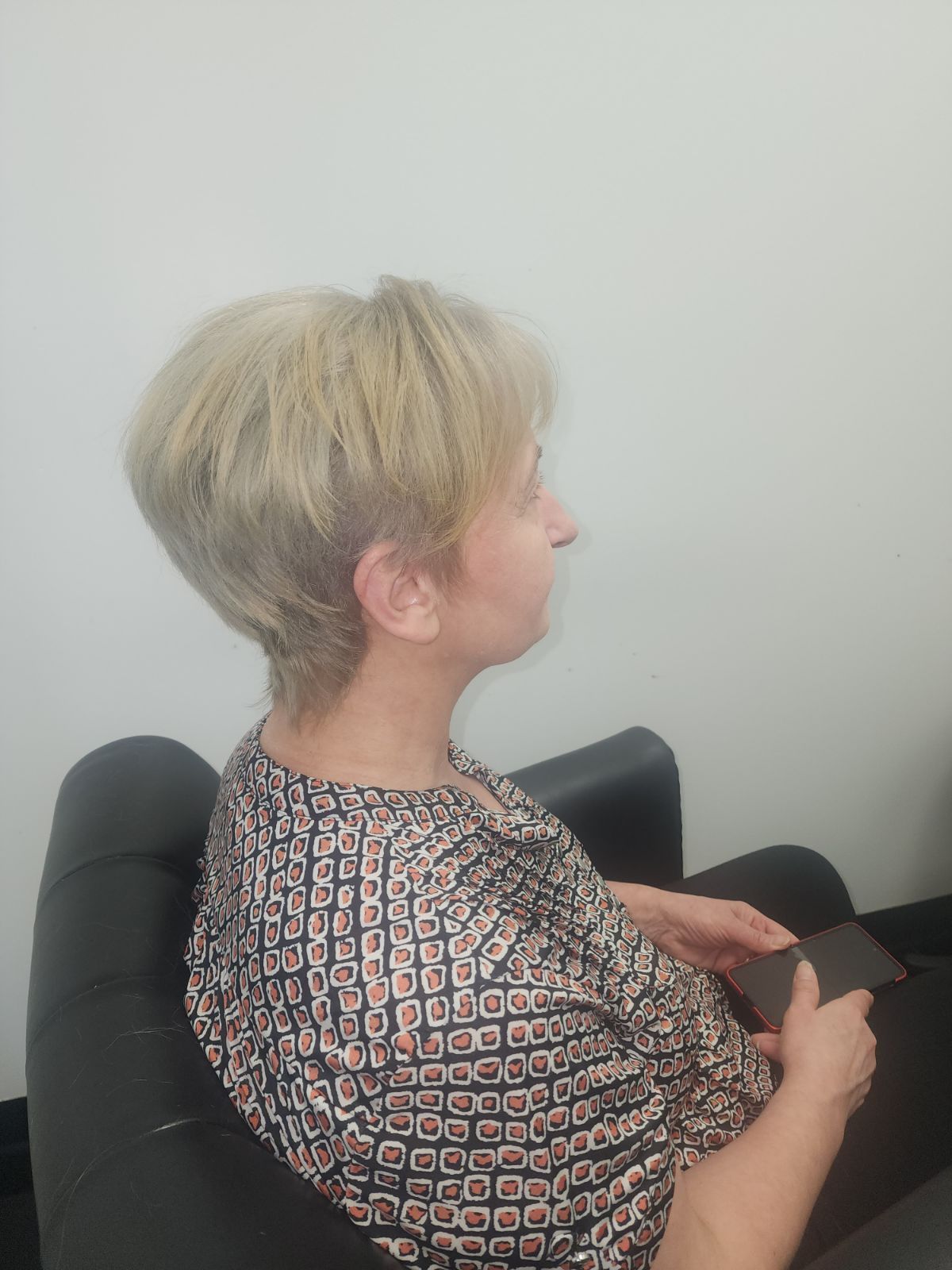 A woman with short blonde hair is sitting in a chair holding a cell phone.