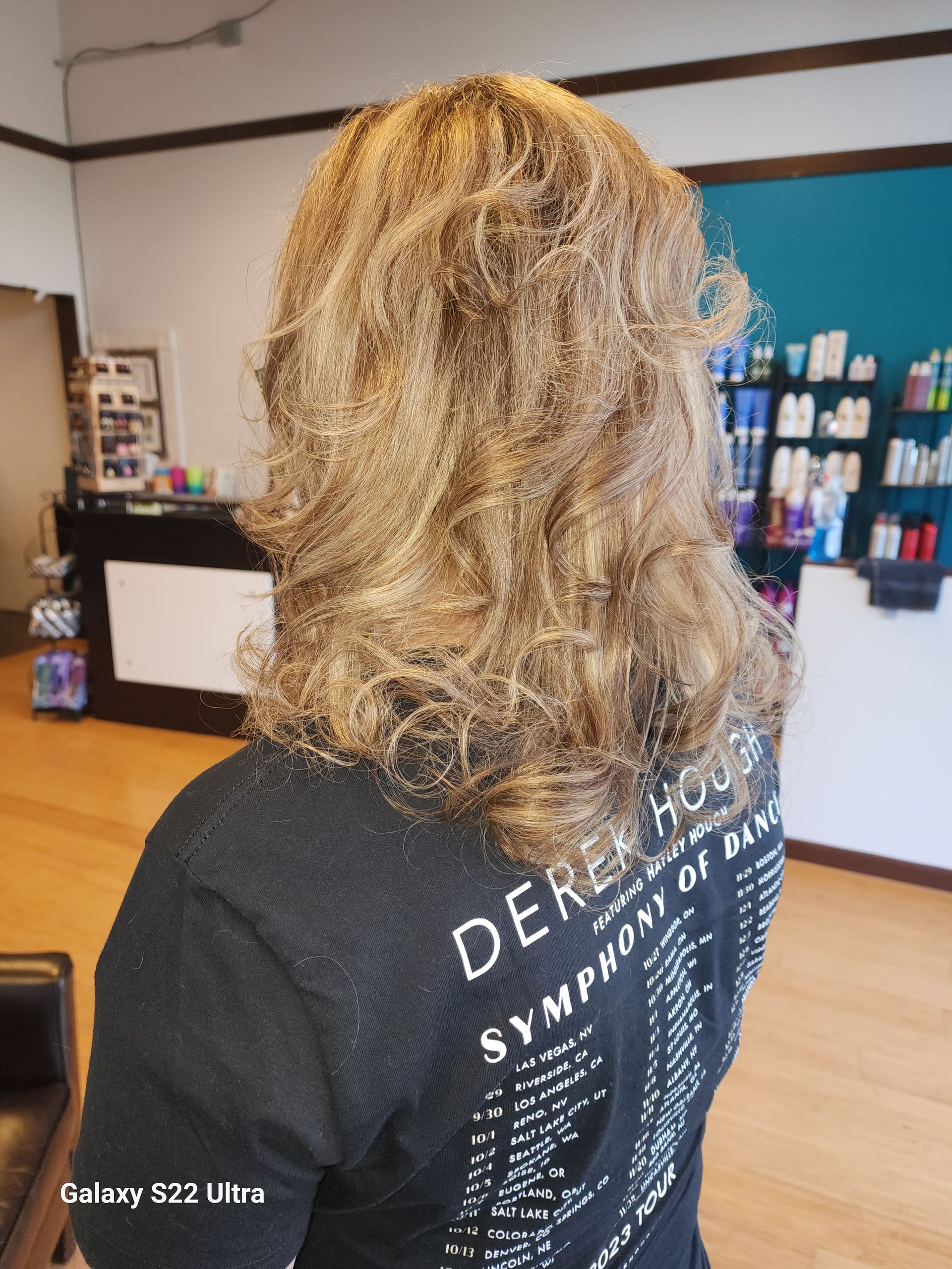 A woman with long blonde hair is wearing a black shirt with the word derek house on it.