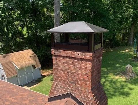 Brick chimney with metal cap beside a small shed in a green wooded yard.