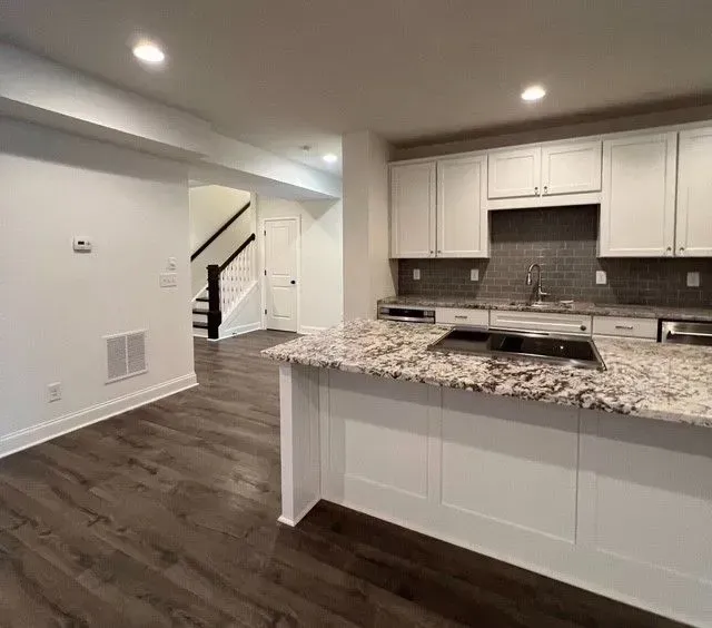 Open kitchen with granite island, white cabinets, dark wood floors, and staircase in the background