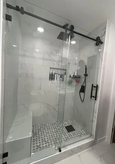 Glass shower enclosure with black fixtures and a white tiled floor in a bright bathroom
