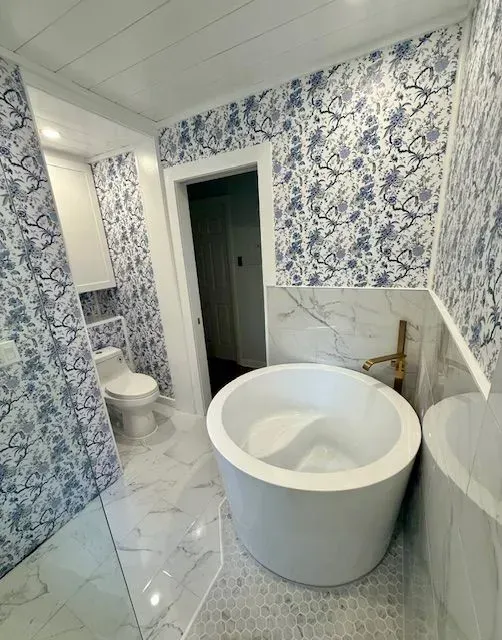 Small bathroom with blue floral wallpaper, white toilet, and freestanding tub near a doorway.