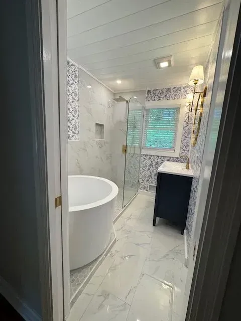 Narrow bathroom with white marble tile, freestanding tub, glass shower, and window with green blinds