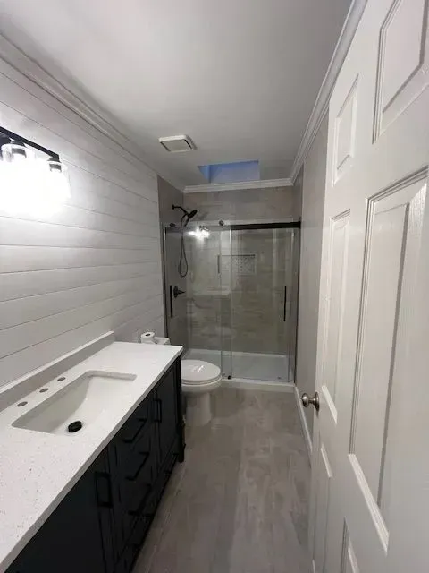 Small bathroom with white vanity, toilet, and glass shower enclosure in a narrow hallway.