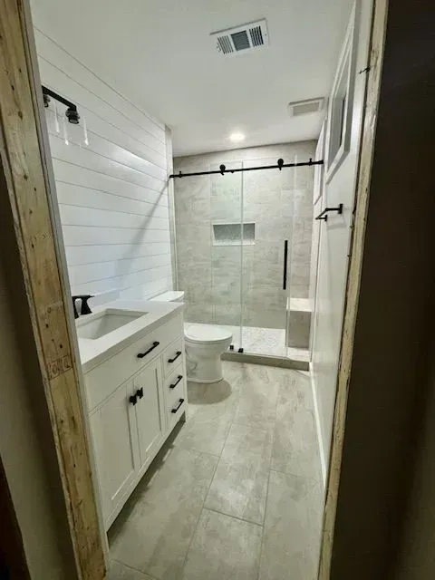 Narrow white bathroom with vanity, toilet, and glass shower enclosure on beige tile floor