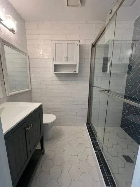 Small white-tiled bathroom with glass shower, vanity sink, toilet, and wall cabinet.