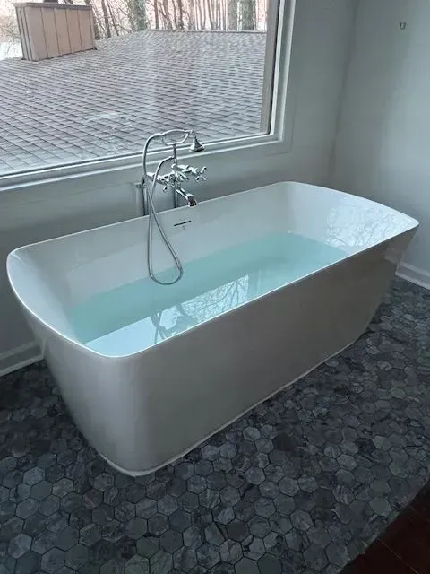 Modern white freestanding bathtub by a large window, filled with water in a tiled bathroom.