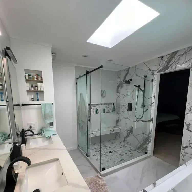 Modern bathroom with glass shower enclosure, white vanity, marble walls, and skylight.