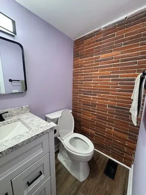 Small bathroom with purple wall, brick accent wall, white vanity, toilet, and towel hanging nearby.