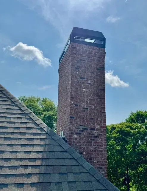Brick chimney tower beside a shingled roof under a blue sky with clouds