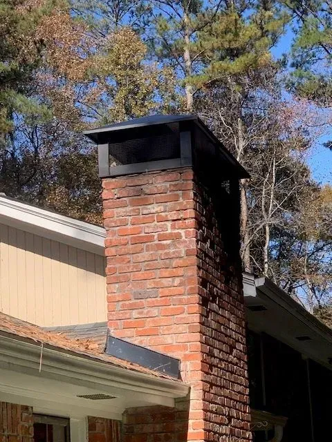 Red brick chimney with black metal cap on a house roof against autumn trees and blue sky