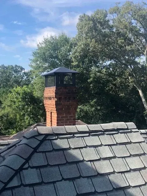 Brick chimney on a gray shingled roof beneath blue sky and leafy trees