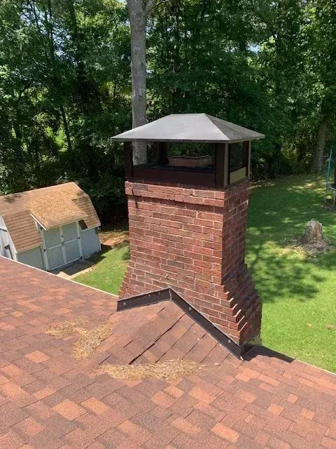 Brick chimney with metal cap on a shingled roof beside a small shed in a wooded yard