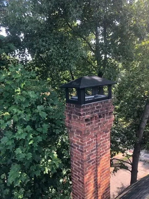 Brick chimney with black cap rising above dense green trees