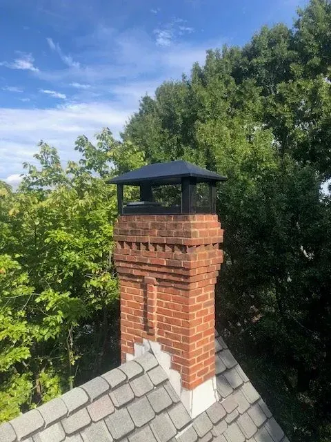 Red brick chimney with black cap above gray shingle roof, surrounded by green trees and blue sky