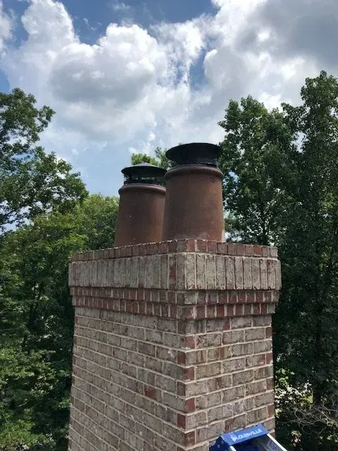 Brick chimney with two metal flues rising above green trees under a cloudy sky