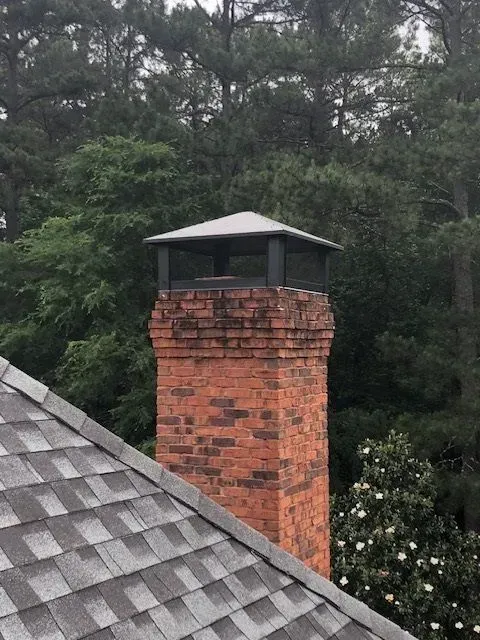 Brick chimney with a metal cap beside a gray shingled roof, backed by dense green trees