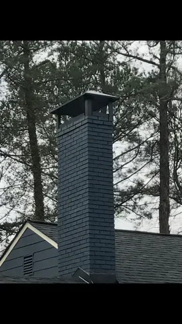Blue-gray brick chimney on a house roof with a cap, set against tall trees.