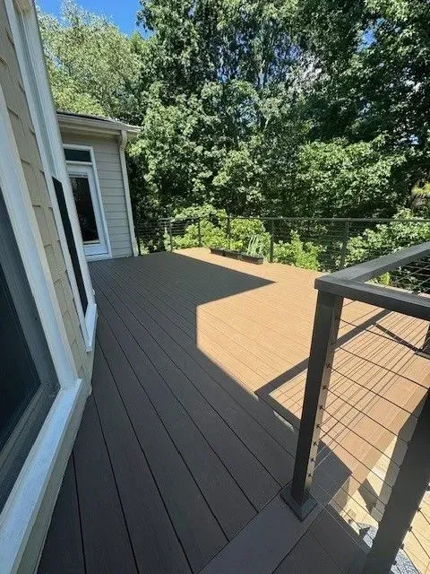 Wooden deck with railing beside a house, surrounded by trees and bright sunlight.
