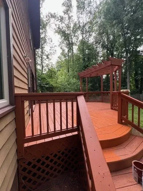 Wooden deck with red railings and steps, extending to a pergola among trees beside a house