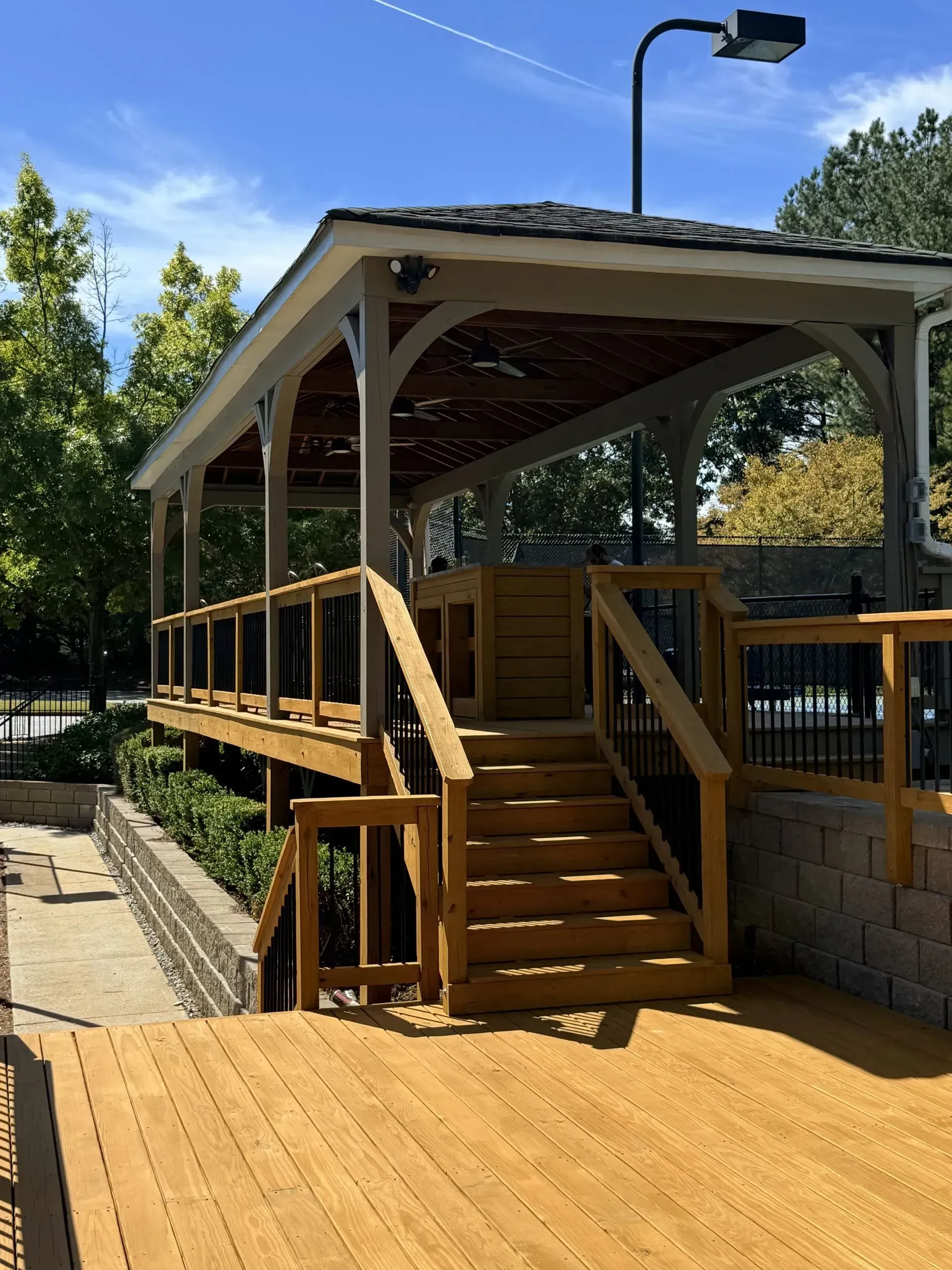 Wooden outdoor gazebo with stairs and railing beside a paved walkway, under a clear blue sky.