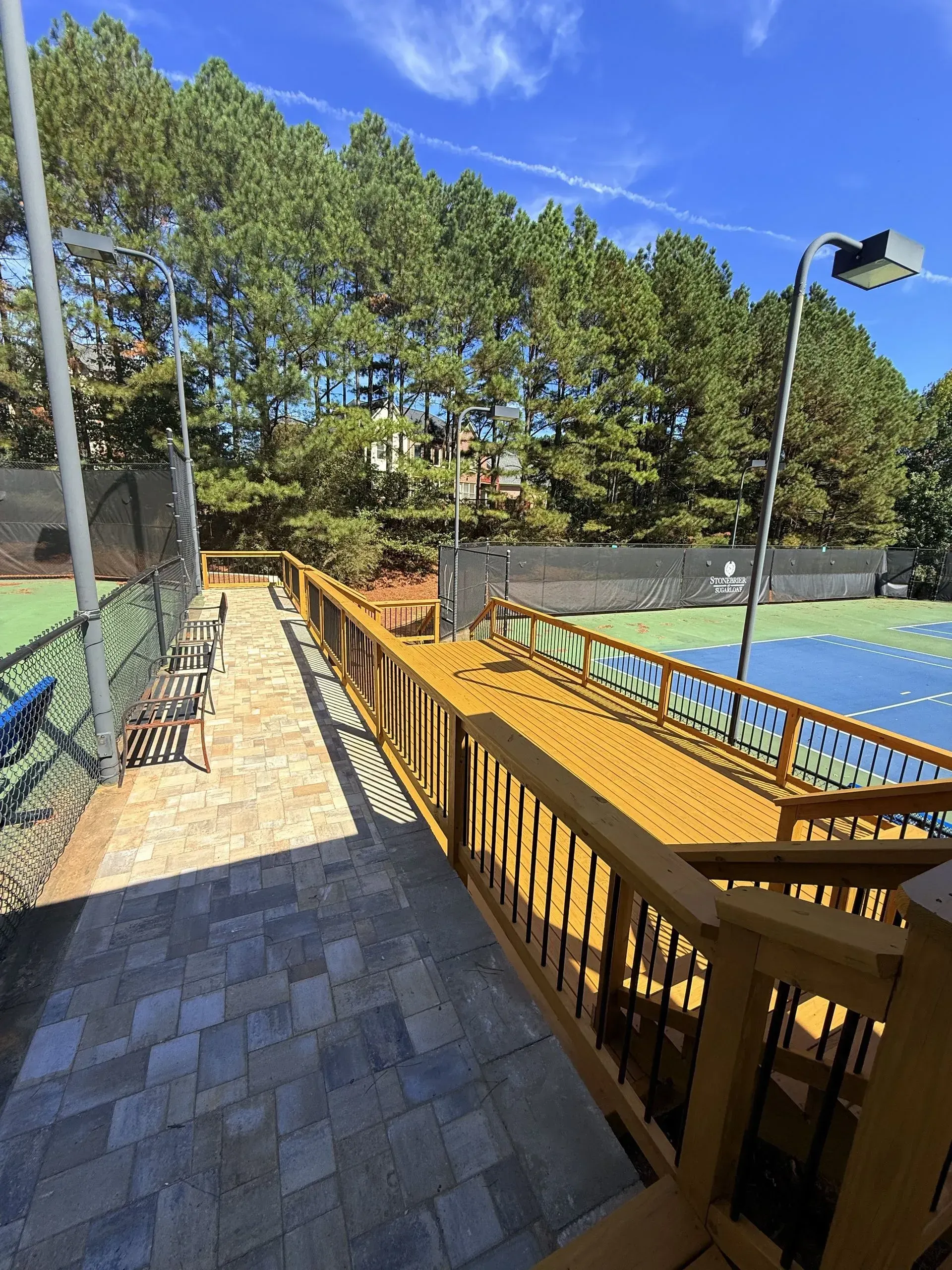 Outdoor walkway beside a blue tennis court, with a wooden ramp and benches under a clear sky