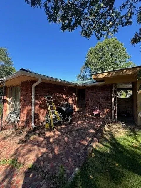 Brick house patio with a ladder, motorcycle, and shaded yard under a bright blue sky