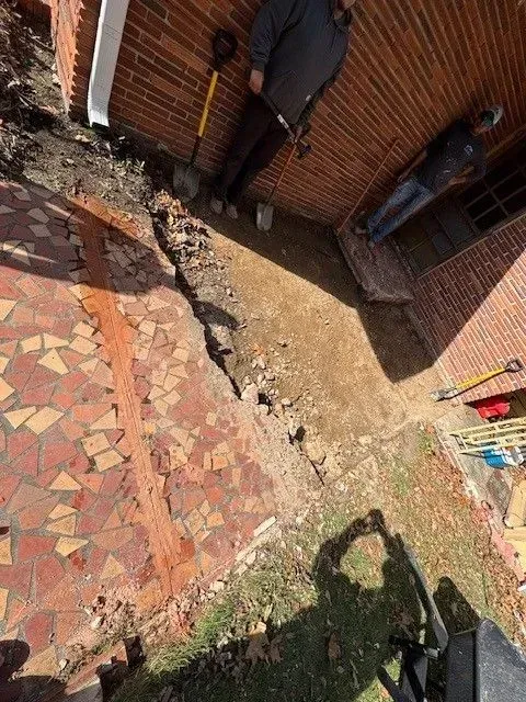 Tilted view of a brick courtyard with a stone path, black gate, and a person’s shadow on the grass