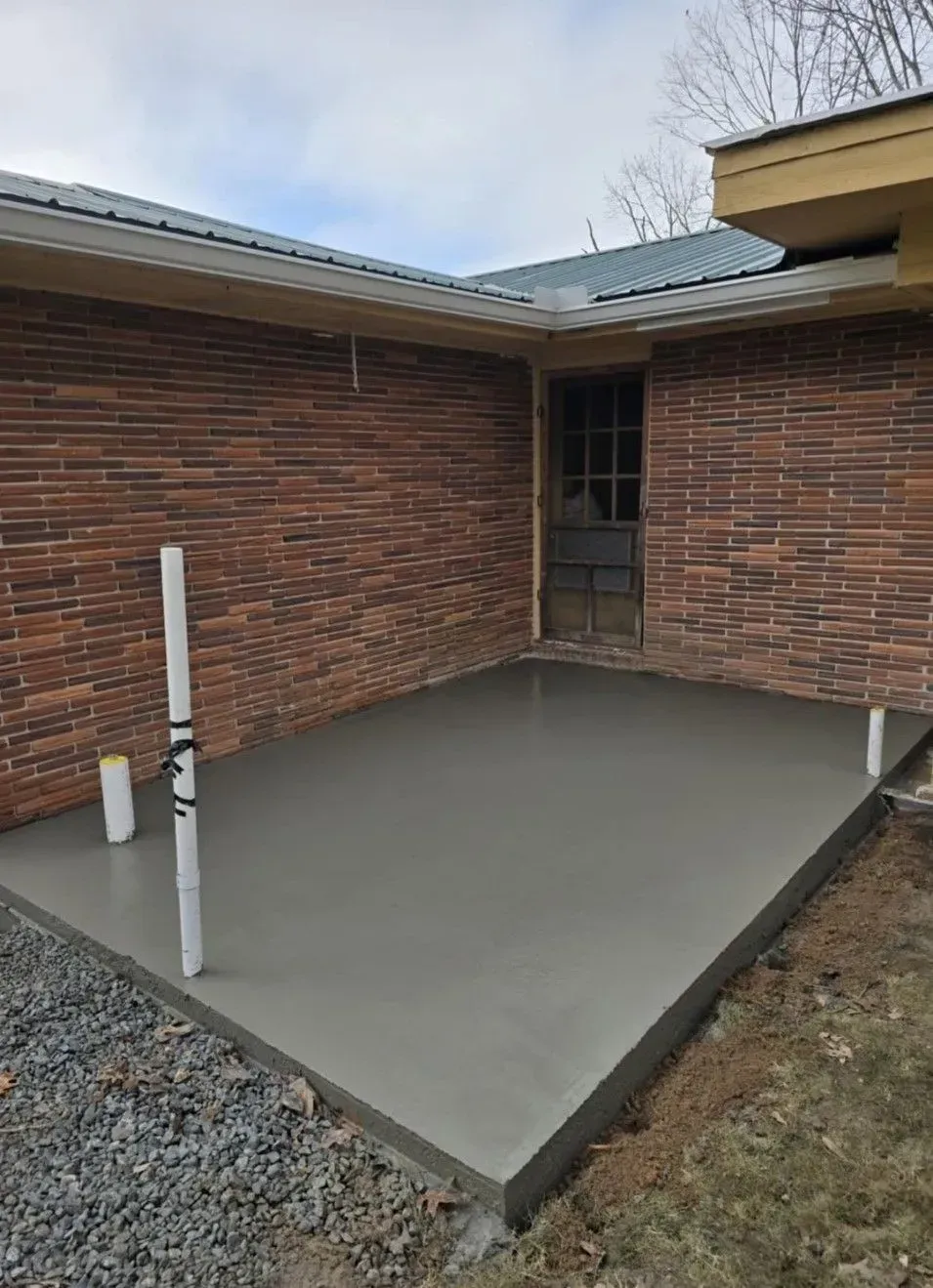 Freshly poured concrete patio beside a brick house, with a white pipe and gravel edging.