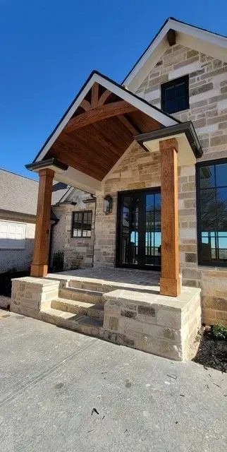 Stone house entrance with wooden porch roof, glass double doors, and two wooden posts on a paved driveway