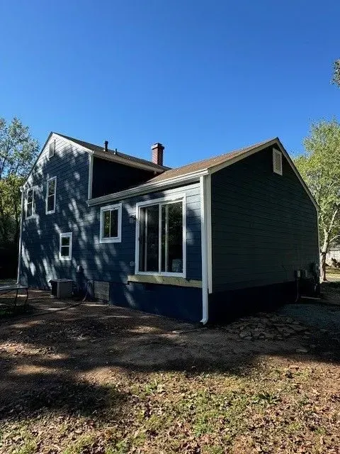 Blue house with white trim and large windows under a clear sky, surrounded by trees and yard.