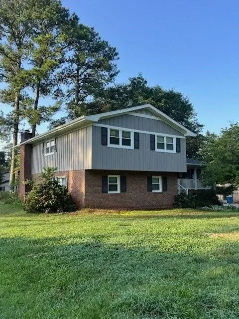 Two-story house with brick and gray siding on a grassy lot, surrounded by tall trees under a blue sky