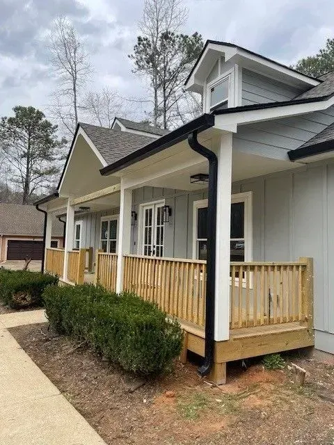 Covered porch with wooden railings on a light-colored house beside a sidewalk and shrubs