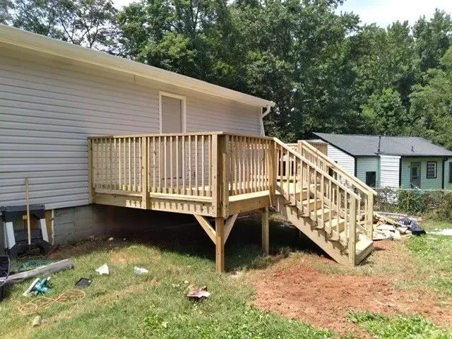 Wooden deck and staircase attached to a white house in a grassy backyard, with trees and a small shed nearby