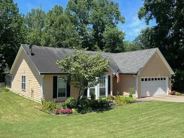 Suburban ranch house with tan siding, gray roof, and a garage, surrounded by green lawn and trees