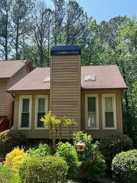 Suburban house with tan siding, brown roof, central chimney, and green shrubs in front of tall trees