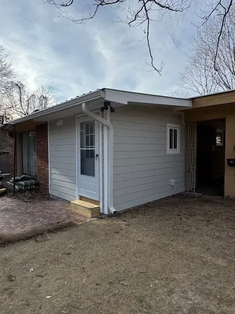 Small white house exterior with a covered carport and a front door beside a brick wall.