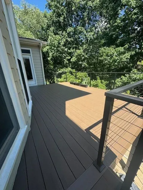 Wooden deck beside a house, with black railing and trees in bright daylight.
