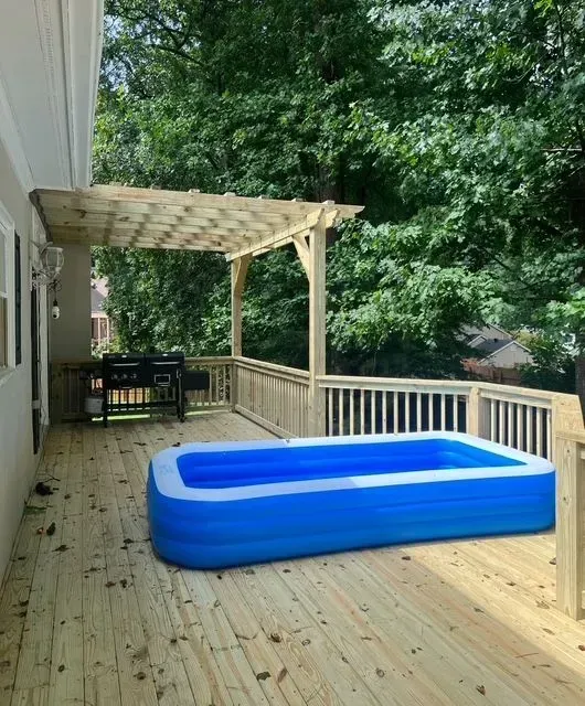 Back deck with a blue inflatable kiddie pool and wooden pergola, bordered by trees.