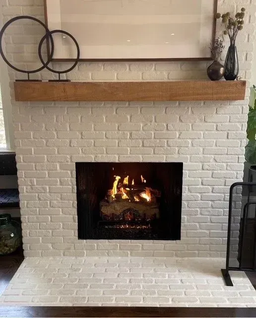 White brick fireplace with a lit fire, wood mantel, decorative rings, and vases in a living room.