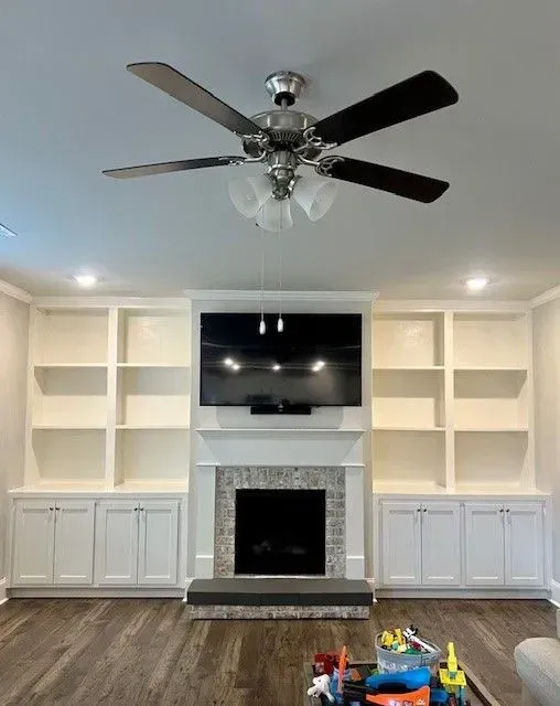 Living room with built-in white shelving, fireplace, mounted TV, and dark ceiling fan on wood flooring