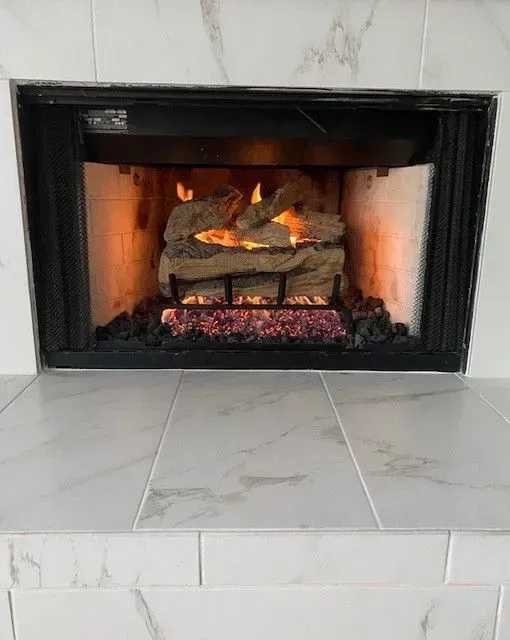 Lit gas fireplace with stacked logs and pink embers in a white tiled surround.