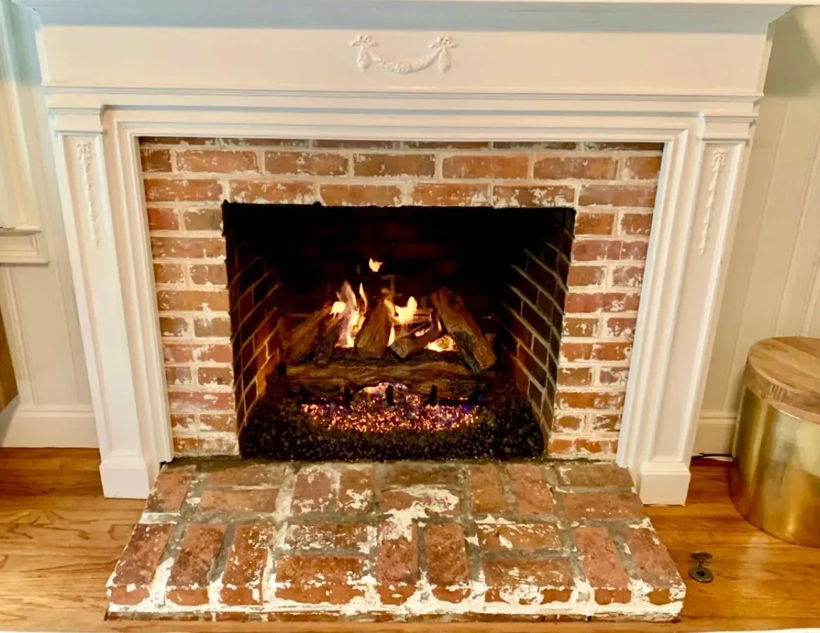 Lit brick fireplace with a burning fire, white mantel, and worn brick hearth in a cozy room