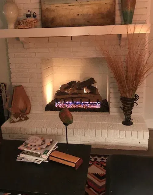 Cozy fireplace nook with glowing electric fire, white brick mantel, decorative vases, and magazines on a table