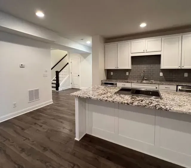 Modern kitchen with white cabinets, granite island, dark wood floors, and a staircase visible in the background