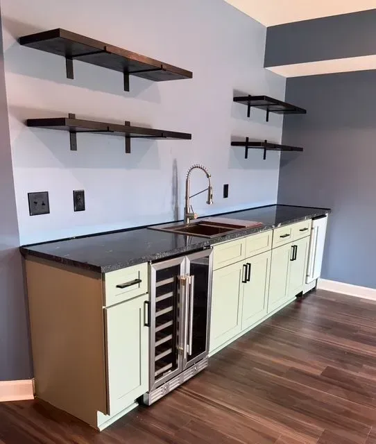 Modern kitchen with pale cabinets, black countertops, wall shelves, sink, and a wine fridge.