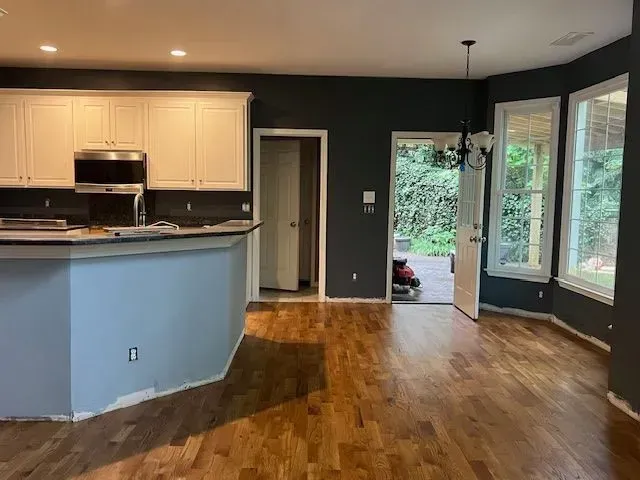 Empty modern kitchen and living area with hardwood floors, gray walls, and large windows overlooking greenery